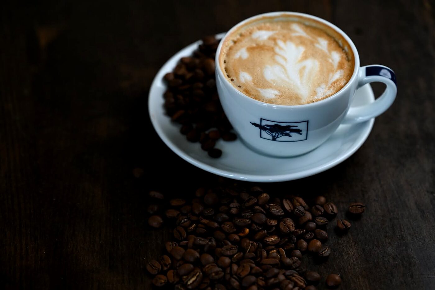 Cappuccino cu latte art într-o ceașcă albă, pe lemn închis, lângă boabe de cafea
