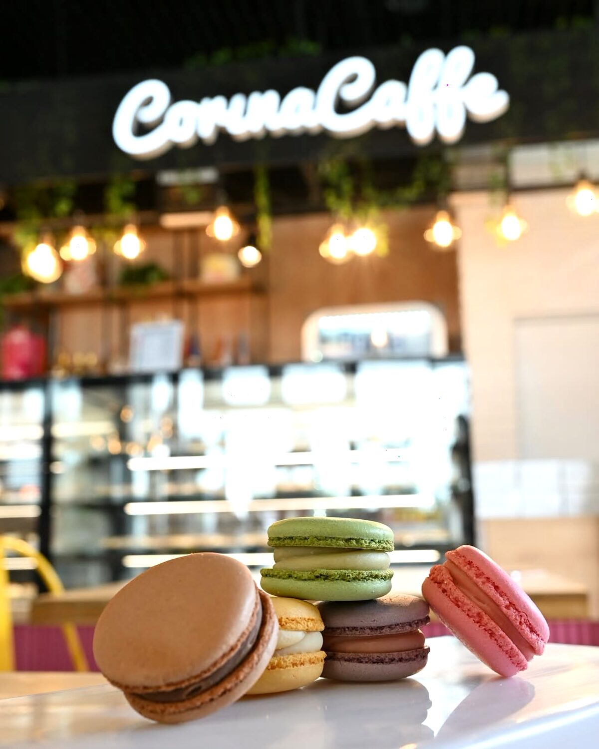 Macarons colorați în prim-plan, cu vitrina Corina Caffe neclară în spate